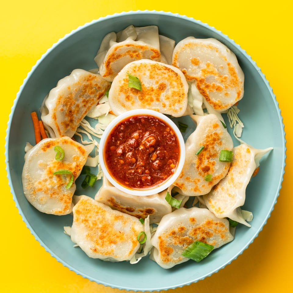 Pan-fried Momo House dumplings with spicy red chili sauce, cabbage, and carrots in a blue bowl.