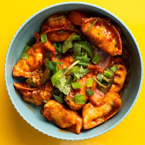A blue bowl filled with chili momos, garnished with green onions and cilantro, on a yellow background.